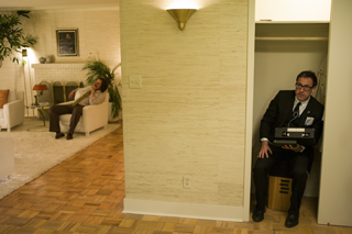 A man in a suit sits in a doorway while another man rests on a white sofa in a living room.