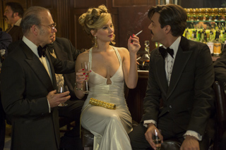 A woman in a white dress sits between two men in tuxedos at a bar, holding a drink.