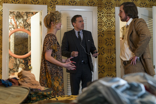 A woman in a floral dress and a man in a suit talk to a man in a brown blazer in a patterned room.