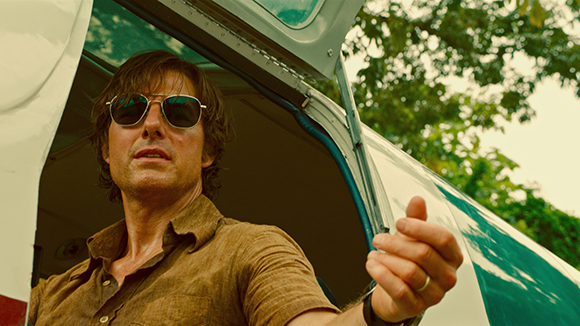 A man wearing sunglasses and a brown shirt looks out from the open door of a white vehicle.