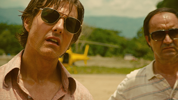 Two men wearing sunglasses stand outdoors near a yellow helicopter in a sunny, tropical setting.
