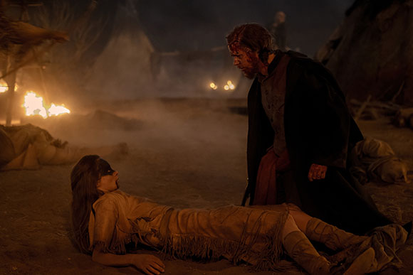 A bloodied man stands over a woman lying motionless on the ground in a dark, smoky landscape.