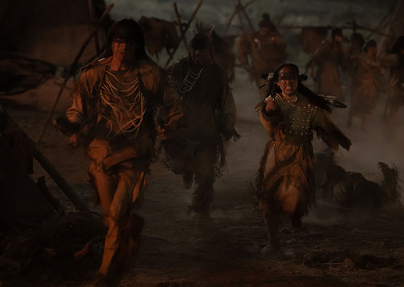 Several people in traditional indigenous clothing run through a dark, dusty landscape.