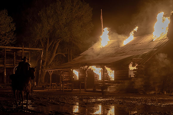 A person on horseback watches a wooden building burn with large flames and smoke at night.