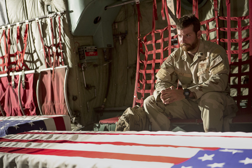 A bearded soldier in desert camouflage sits inside a cargo plane near an American flag.