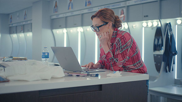 A woman with glasses and red hair works on a laptop in a brightly lit locker room.