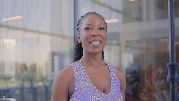 A smiling woman with braided hair wearing a purple sleeveless dress with silver sequins.