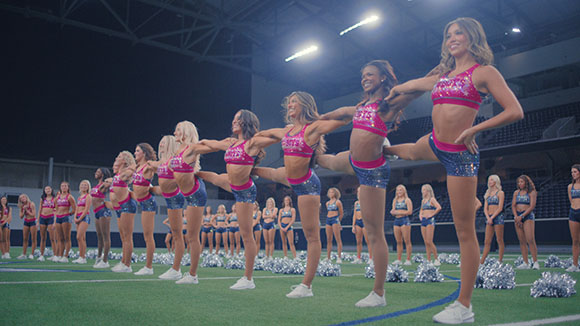 A line of cheerleaders in pink and blue uniforms posing with one leg extended on a green field.