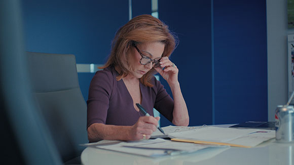 A woman with reddish-brown hair and glasses sits at a desk, writing on documents with a pen.
