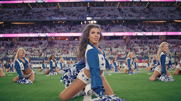 A group of cheerleaders in blue and white uniforms performing on a green football field in a large stadium.