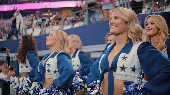 Cheerleaders in blue and white star-patterned uniforms smiling in a stadium.