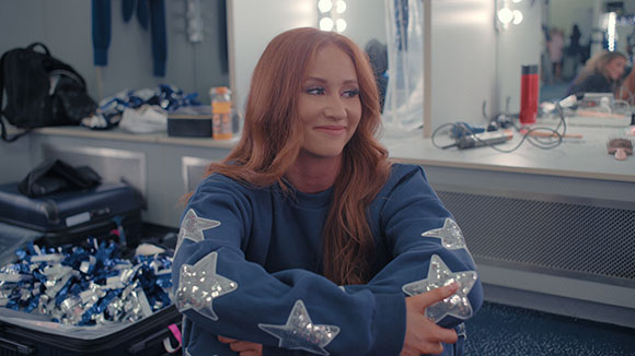 A smiling woman with long wavy red hair wearing a navy blue sweatshirt with silver stars.