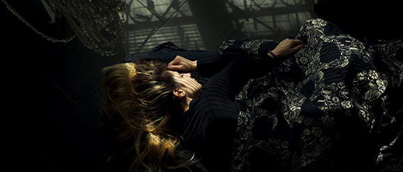 A person with long blonde hair lies on a dark, floral patterned fabric in a dimly lit room.