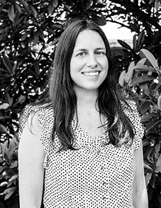 A smiling woman with long dark hair wearing a polka dot sleeveless top, standing before leafy bushes.