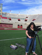 A woman in a black shirt and jeans operates a professional video camera on a tripod on a green football field.