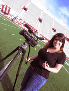 A smiling woman in a black shirt and jeans stands next to a video camera on a tripod on a green sports field.