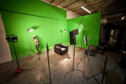 A large green screen studio filled with various lighting rigs, tripods, and a person in high-visibility gear.