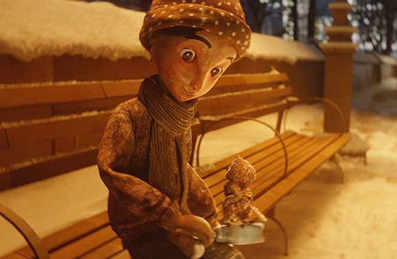 A small puppet wearing a scarf and polka-dot hat sits on a wooden bench in a snowy park.