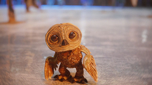 A small, brown, stylized owl figurine with large eyes standing on a light-colored reflective floor.