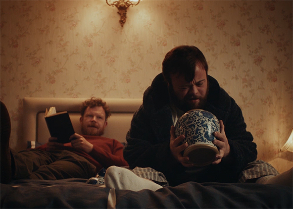 Two men sit on a bed in a room with floral wallpaper; one holds a book, the other holds a blue and white jar.