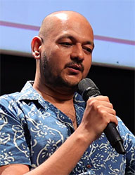 A bald man in a blue patterned shirt speaks into a black handheld microphone.