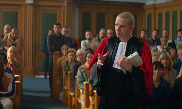 A man in black and red legal robes gestures while speaking in a courtroom filled with spectators.