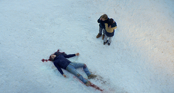 A person lies in the snow near two people hugging in a wide snowy landscape.