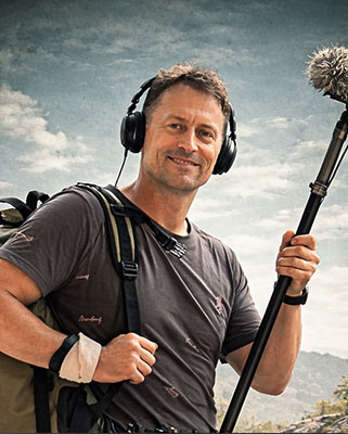 A man with a backpack, headphones, and a boom microphone smiling against a cloudy sky.