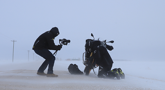 A person in dark clothing films a motorcyclist kneeling in a heavy snowstorm on a flat landscape.
