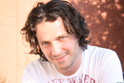 A man with wavy dark hair and blue eyes smiling while wearing a white t-shirt.