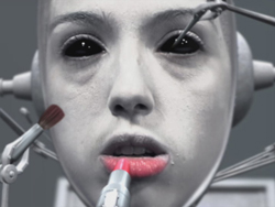A pale humanoid robot face being touched by mechanical tools applying lipstick and eye makeup.