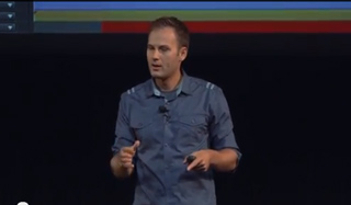 A man in a blue button-down shirt speaking on a stage against a dark background.