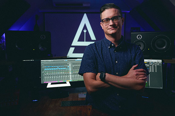 A man with glasses and a blue patterned shirt stands with arms crossed in a music studio.