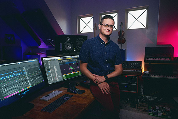 A man in a blue shirt stands in a studio filled with monitors, speakers, and musical instruments.