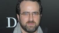 A man with dark hair, glasses, and a beard looking forward against a dark background.