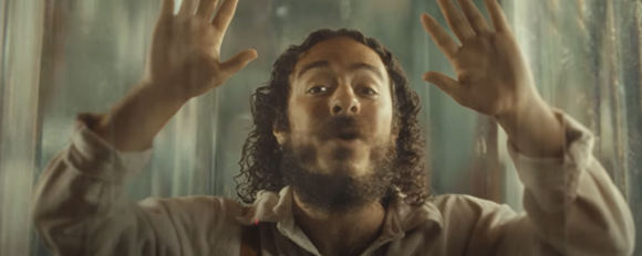 A man with dark curly hair and a beard presses both palms against a transparent glass pane.