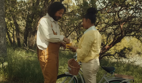 A man in tan overalls and a woman in a yellow shirt stand together in a sunlit forest near a bicycle.