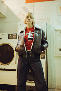 A person in a shearling jacket, red t-shirt, and dark jeans stands in front of washing machines.