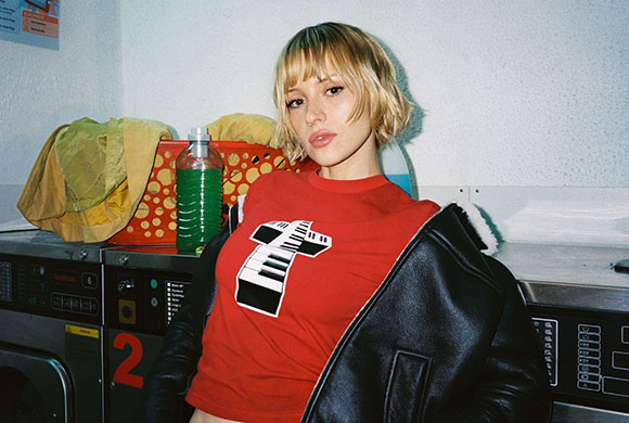 A blonde woman in a red graphic t-shirt and black leather jacket leans against laundry machines.