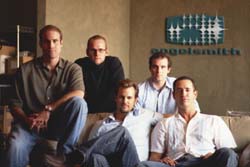 Five men in casual attire posing together in an office with a logo on the wall behind them.