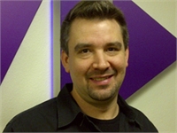 A smiling man with short brown hair wearing a black collared shirt against a purple and white geometric background.