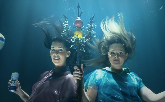 Two women underwater with flowing hair, one holding a trident and the other holding a beverage can.