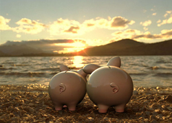 Two ceramic piggy banks sit on a pebble beach facing a sunset over a lake and mountains.