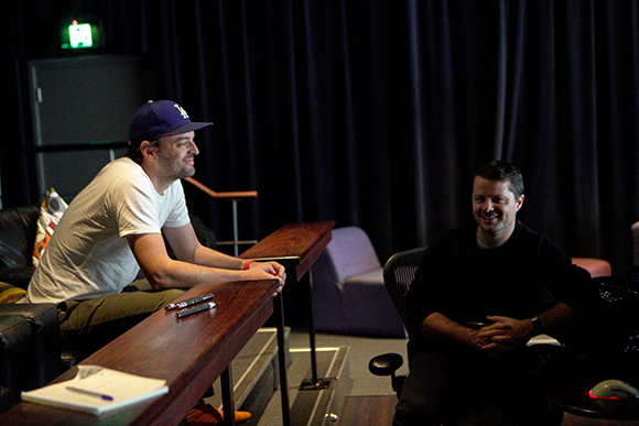 A man in a white t-shirt and blue cap sits at a wooden table talking to a man in a black shirt.