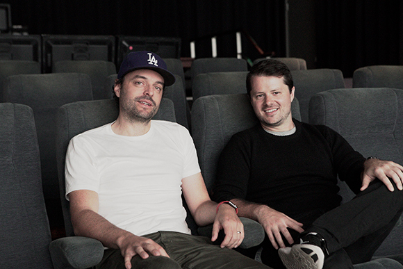 Two men sit side-by-side in gray theater seats, one in a white t-shirt and cap, the other in a black sweater.