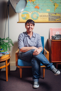 A person with short dark hair sitting in a blue armchair, wearing a grey shirt and jeans.