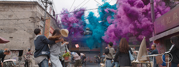 People stand in a narrow street watching large clouds of purple and teal smoke billow overhead.