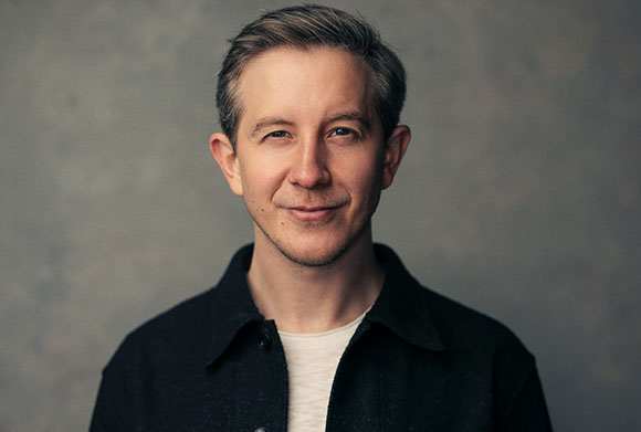 A man with short brown hair and a slight smile wearing a black button-down shirt over a white t-shirt.