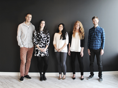 Five adults standing in a line against a dark gray wall on a light wood floor.