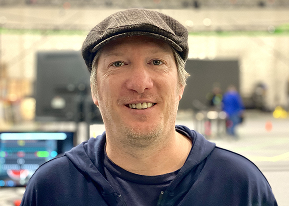 A smiling man wearing a dark flat cap and a navy blue hoodie in a large, blurred indoor venue.